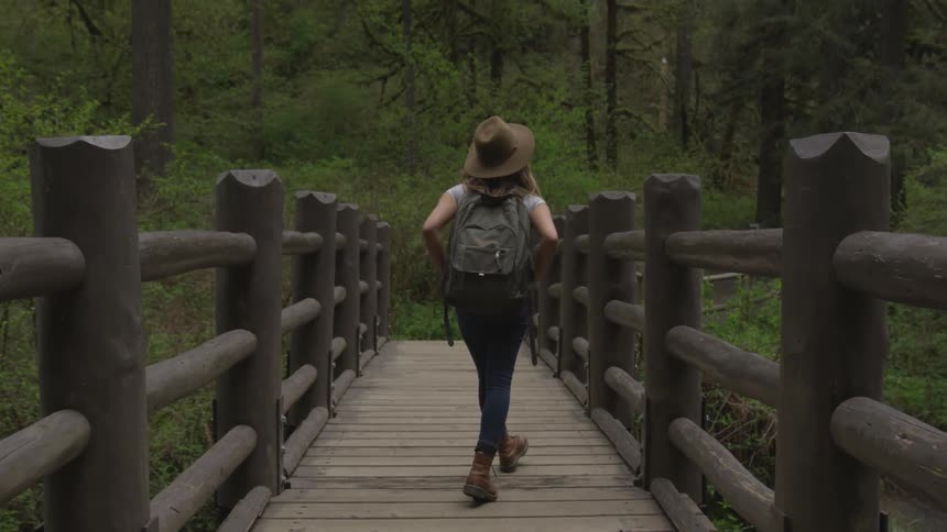 A woman wearing a brown big brimmed hat and a backpack is walking on a wooden bridge in a natural setting maybe a forest