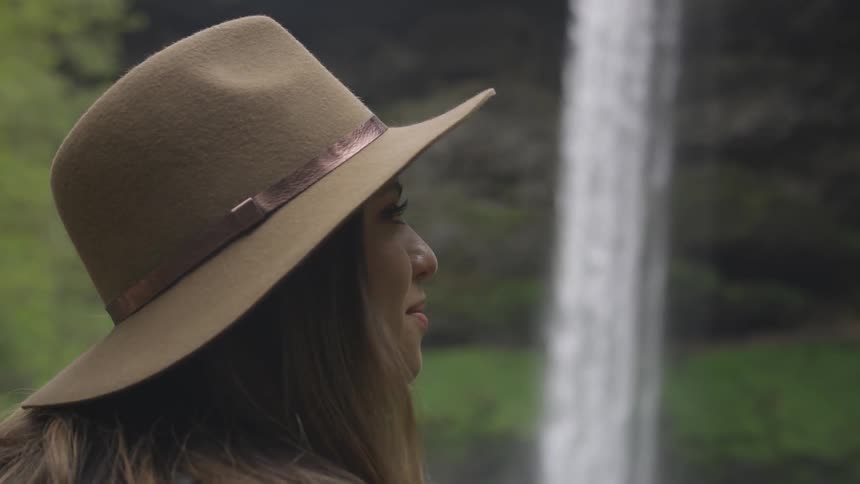 Lady turn right wearing brimmed hat smiles as we see a waterfall in the background