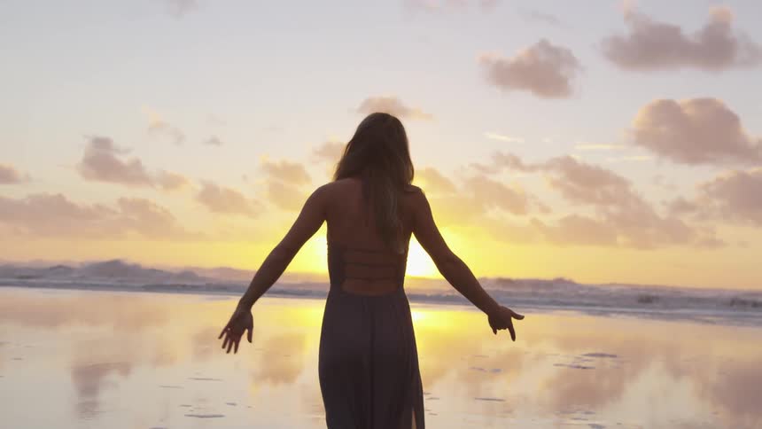 Woman wearing a dress with arms spread out facing the sun setting over the water
