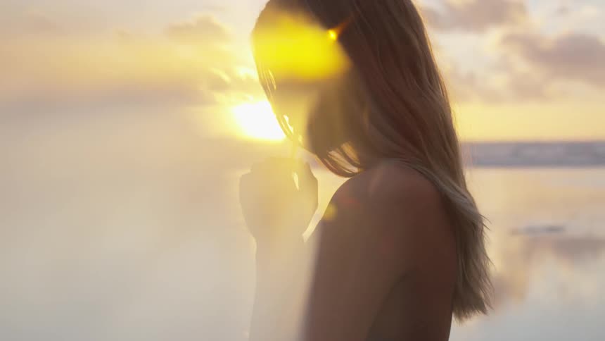 A woman stands near the water with her hands cupped under her chin
