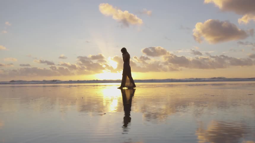Woman walks on wet sand reflecting sunset on the beach