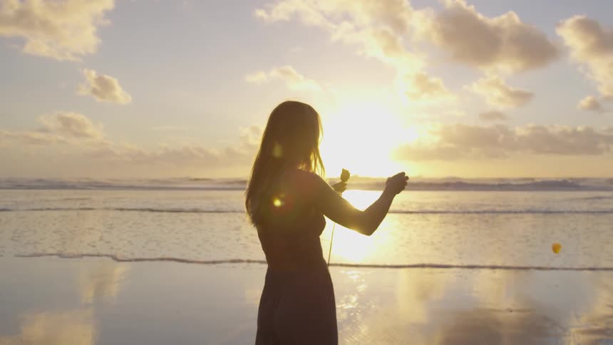 Woman pulls petals off a flower while standing next to the water at sunrise