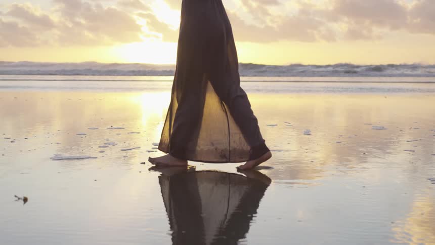 White woman walking on the beach while wearing a long dress during the sunset