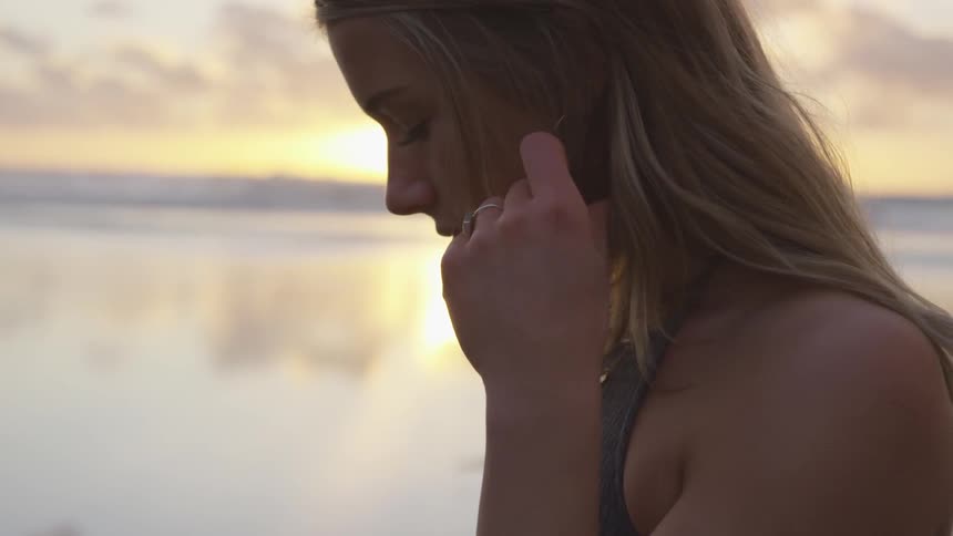 A blonde woman plays with her hair on the beach at sunset next to the ocean