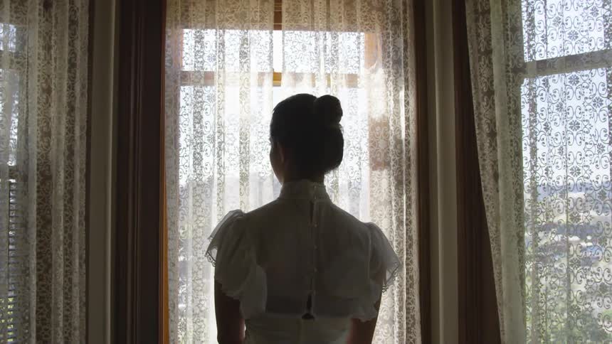 A woman in a white dress standing near a window looking outside