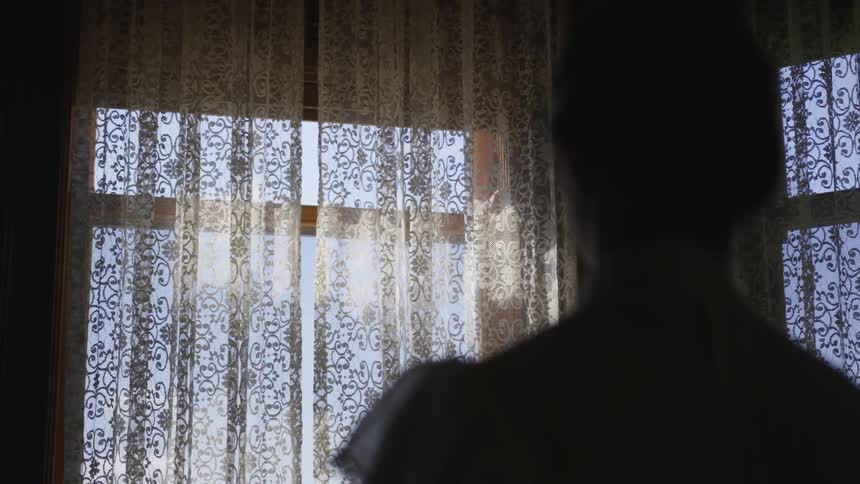 Woman stands in a dim room and looks out window in the daytime