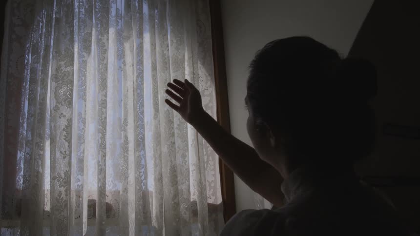 A silhouetted woman raises her hand to a curtained window