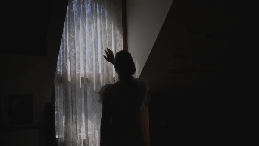 Woman stands in dark room reaching hand toward lit curtained window