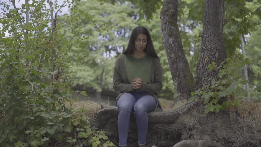 A young woman sits on a tree trunk with her hands clasped together like she is praying