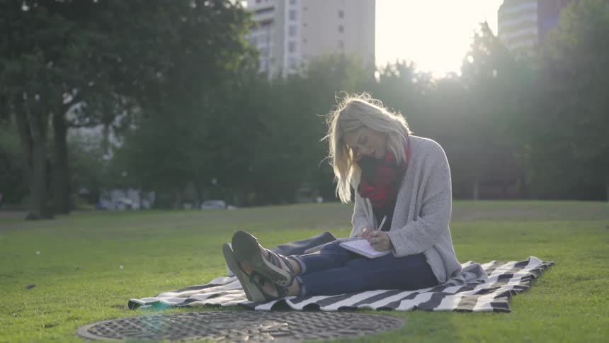 Woman sits on blanket in park writing in journal