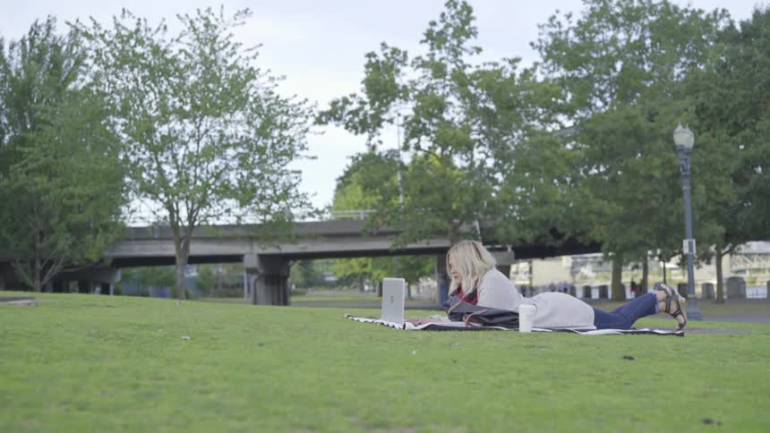 Woman works on her laptop while lying down in a park