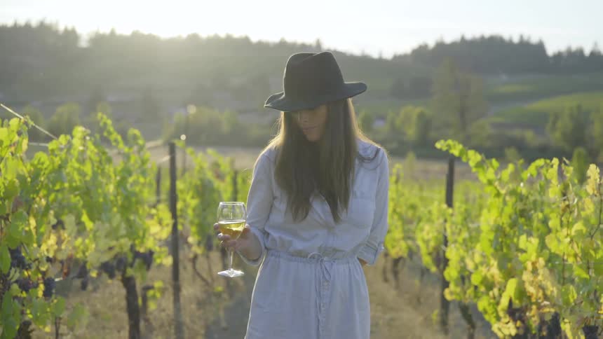 A white woman wearing a hat enjoys a glass of wine in a vineyard