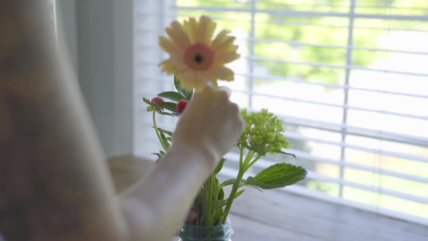 A person arranges flowers in a glass jar near the window