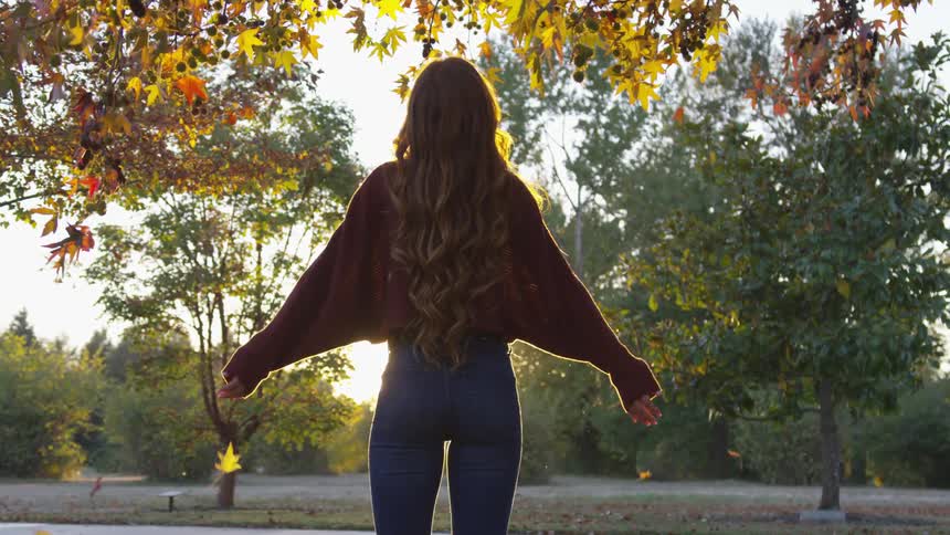 Woman raises her arms under falling leaves with the sun setting
