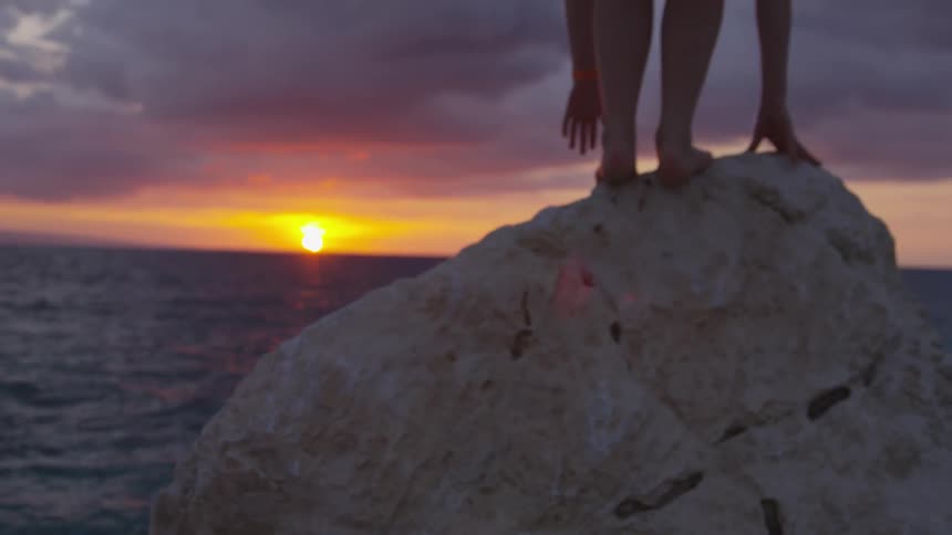 Person climbs and stands on large rock near ocean at dusk