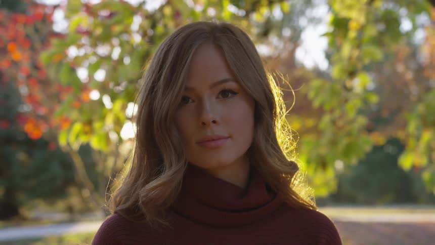 Blonde woman looks ahead with a slight smile under autumn leaves when the sun goes down