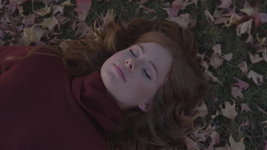 A ginger white woman with her eyes shut lies on the ground covered with fallen leaves