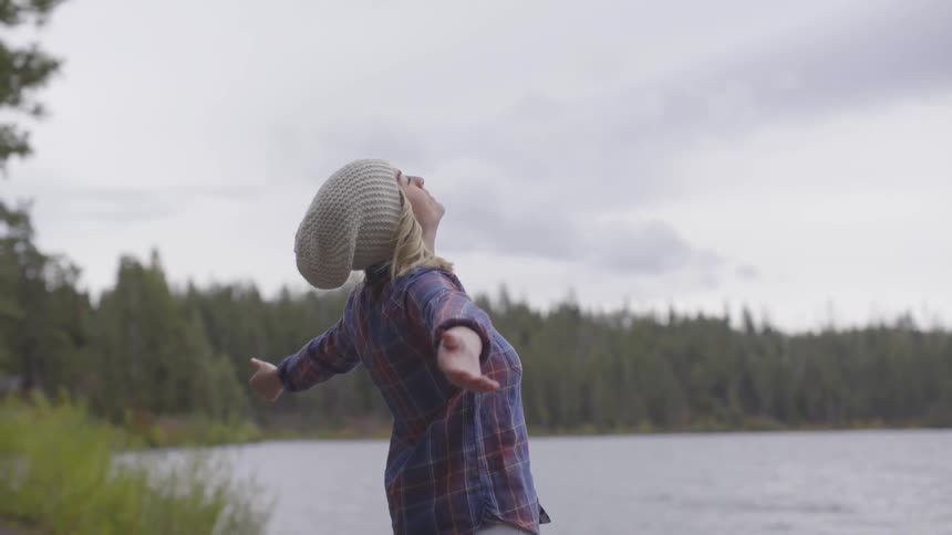 A young woman stands by a lake and extends her arms