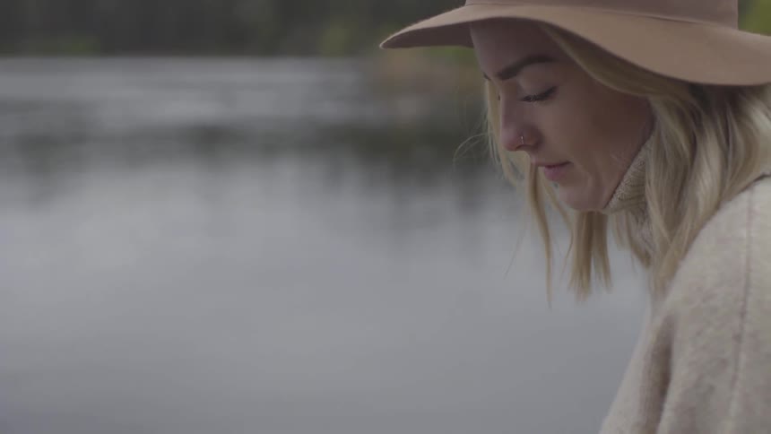 Woman with hat stands near lake and looks down