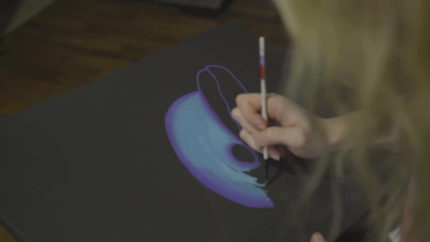 A blonde female artist paints a picture on a black canvas