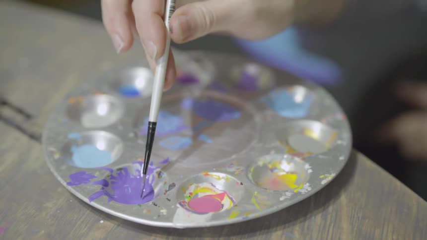 A woman dips an artists paint brush into paint on a palette