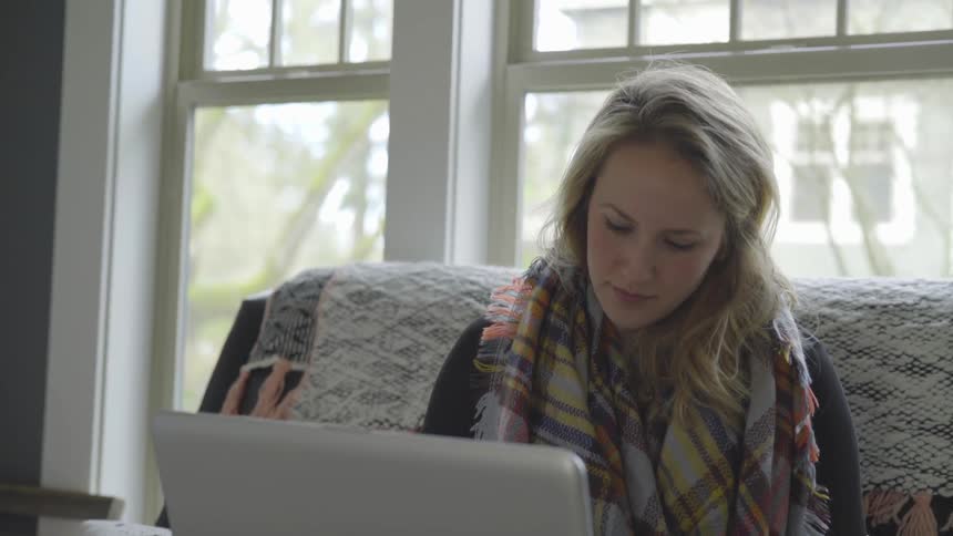 A woman sitting on a couch working on a laptop with windows behind her