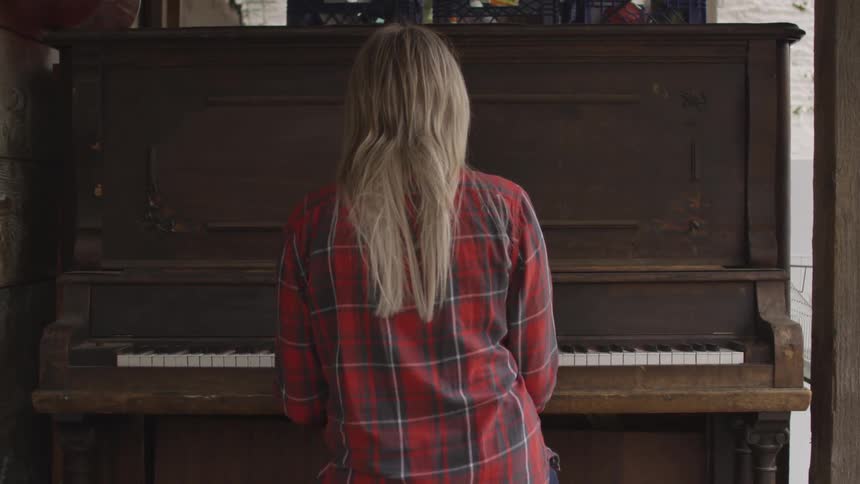A young blonde haired woman in a red flannel shirt sits at an old fashioned wooden piano