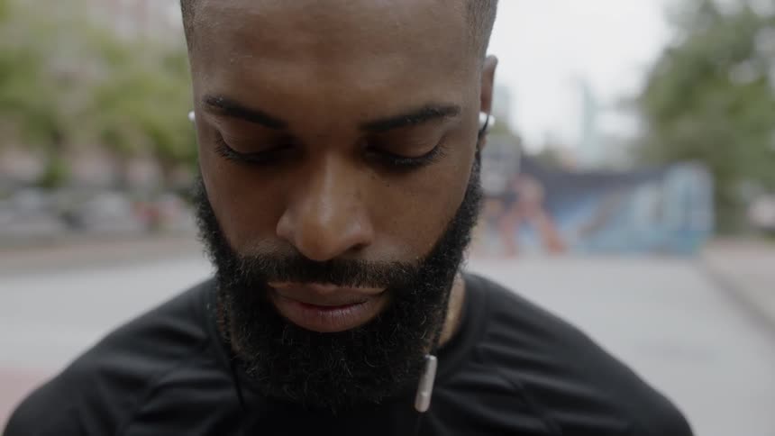 A man wearing earbuds stands on an outdoor court looking down