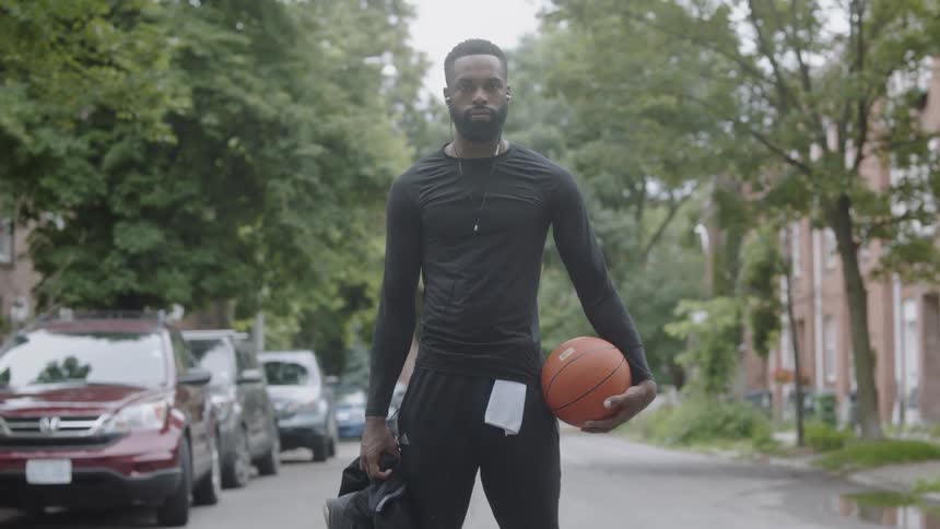 A black man stands in the middle of the street with a basketball at his waist