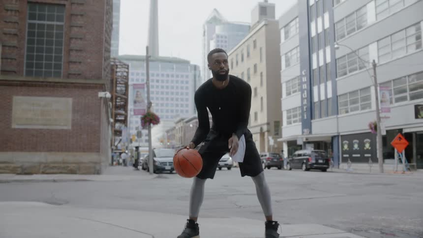 Bearded black man wearing earphones plays basketball on the pavement in the city by day