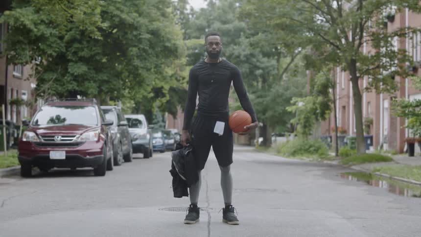 A black man holding a basketball and a backpack stands in a street