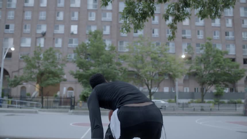 Man sits down on bench and takes off his jacket then standing up holding a basketball in the basketball field