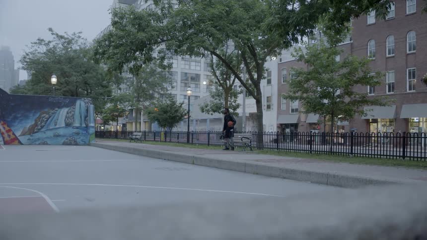 A black man holding a basketball walks down a city sidewalk and sits down on a bench