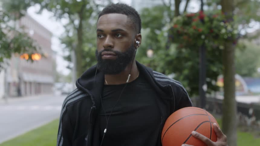 Black man walking down a sidewalk listening to music and holding a basketball