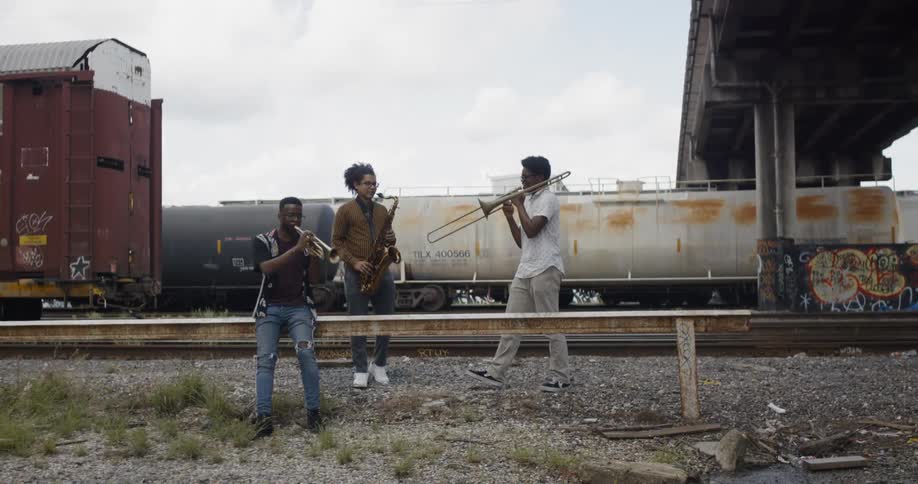 Three men play instruments near a train yard