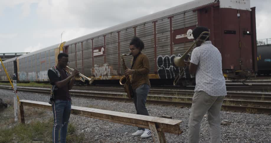 Three black boys play saxophone next to train in the railroad terminal by day