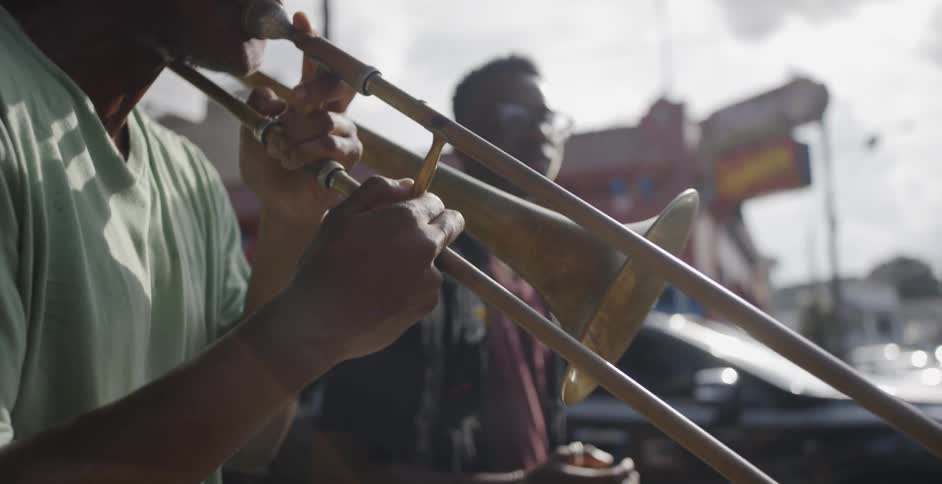 A black man plays the trombone while another black man sways to the music