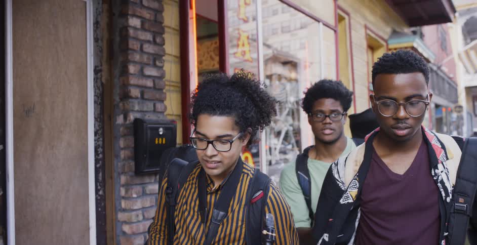 Three black boys wearing glasses and backpack walk down the street and passing the local shops in the daytime