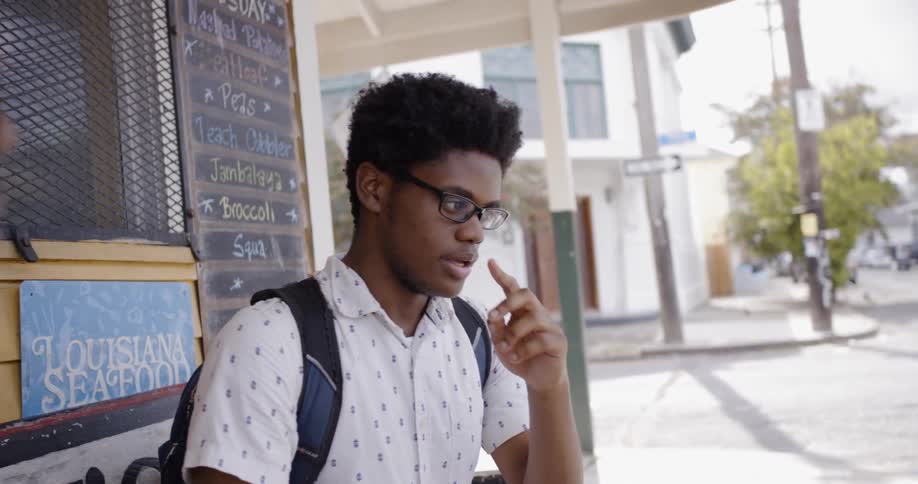Black boy wearing glasses and backpack speaks to another black boy while sitting outside in the daytime