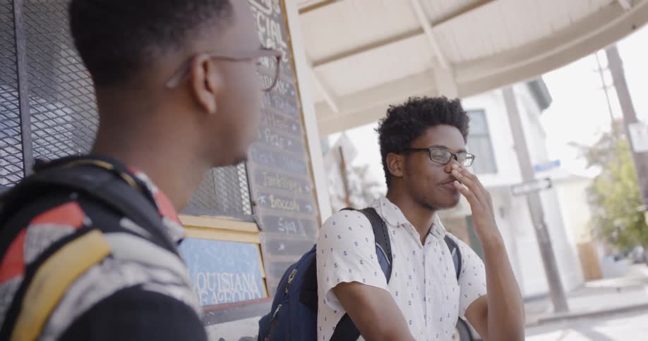 Two young black me sit outside of an outdoor food stand while wearing backpacks and are having a conversation