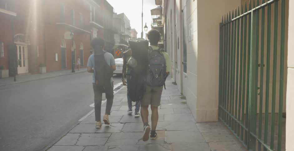Three young man walk down a city street carrying backpacks as well as musical instruments in dark canvas carrying cases