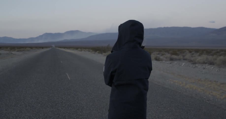Woman in coat stands alone on desert highway at dusk
