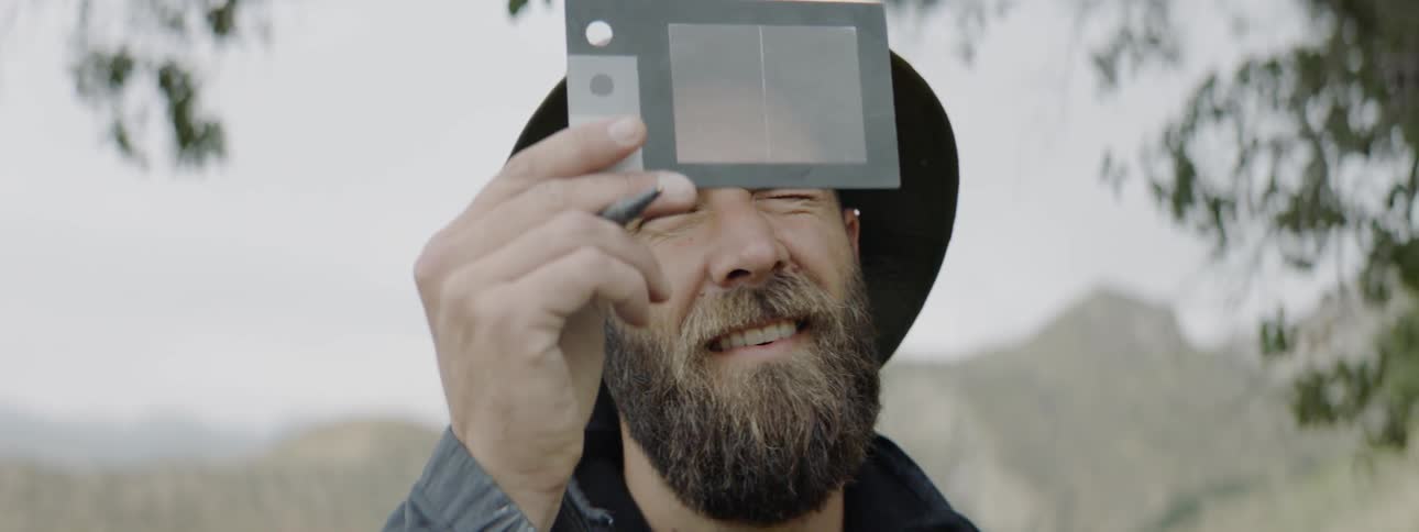 A bearded man in a wearing a hiking hat jacket and backpack lifts a translucent piece of plastic and looks upwards through it while closing his left eye
