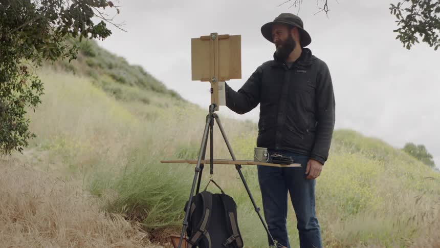 Bearded man squinting and dabbing at a painting on an easel in the outdoors
