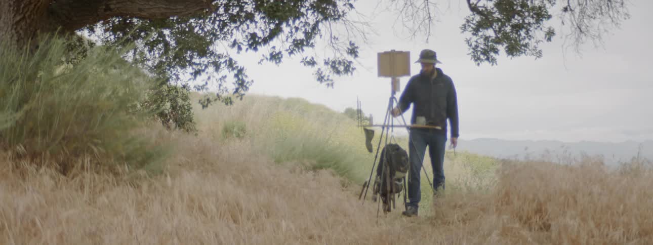 A man stands outside at a portable easel and uses a paint brush