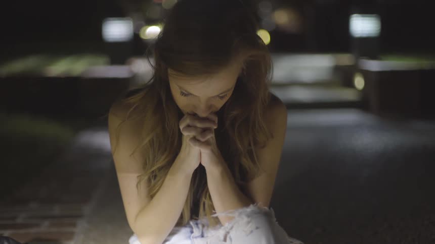 A girl sits in a driveway at night with her hands folded under her face while she looks down