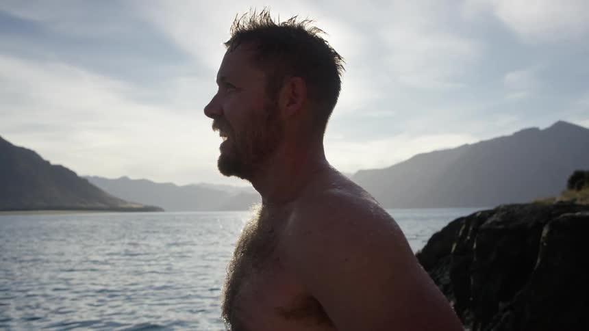 A bearded man stands beside a lake