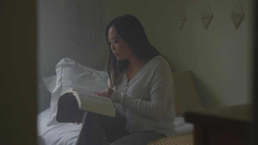 A woman sits on her bed reading a large book which is resting on her knee