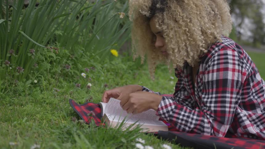 A woman lays on a blanket in the grass reading a book