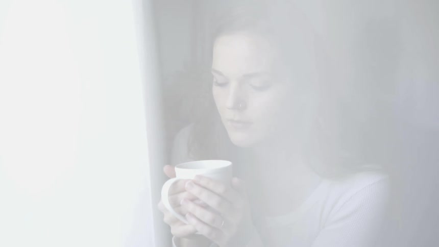 A brunette woman holds a cup by a window